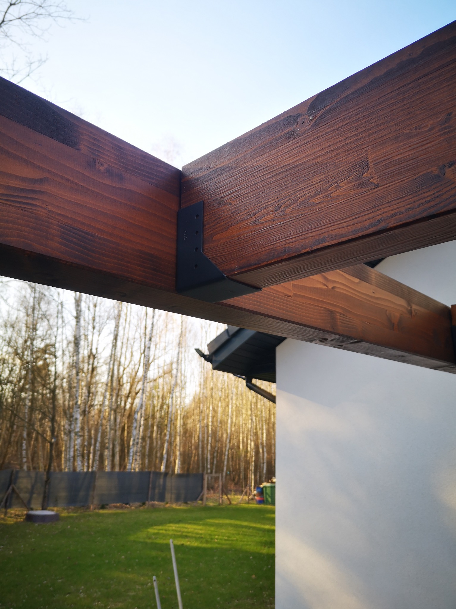 Fragment konstrukcji drewnianej, prawdopodobnie pergoli, z widocznymi elementami mocującymi i tłem z drzew i trawnika w słoneczny dzień. Drewno w ciepłym, brązowym odcieniu.
