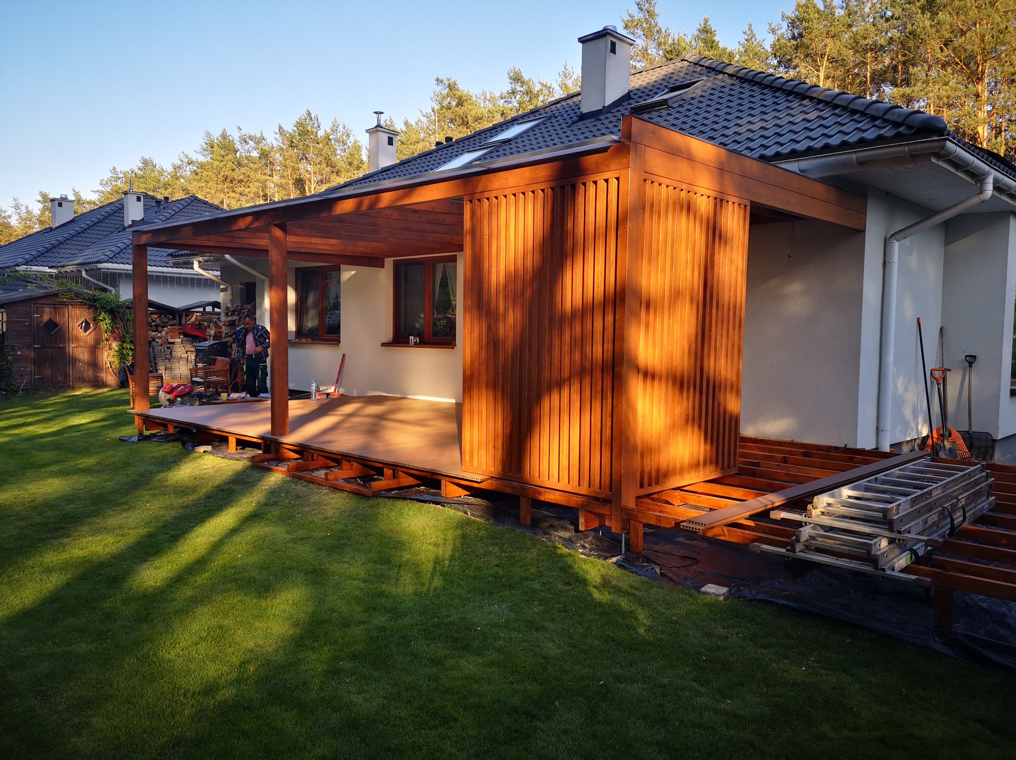 Drewniana konstrukcja tarasu z elementami zabudowy pionowej, widoczna w trakcie montażu. Na pierwszym planie trawnik, w tle dom jednorodzinny i drzewa.