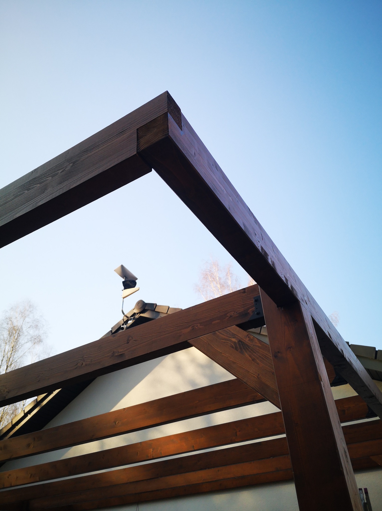 Drewniana pergola z ciemnego drewna, widok z dołu na tle błękitnego nieba. Konstrukcja częściowo zadaszona, widoczny fragment dachu budynku. Mocne słońce.