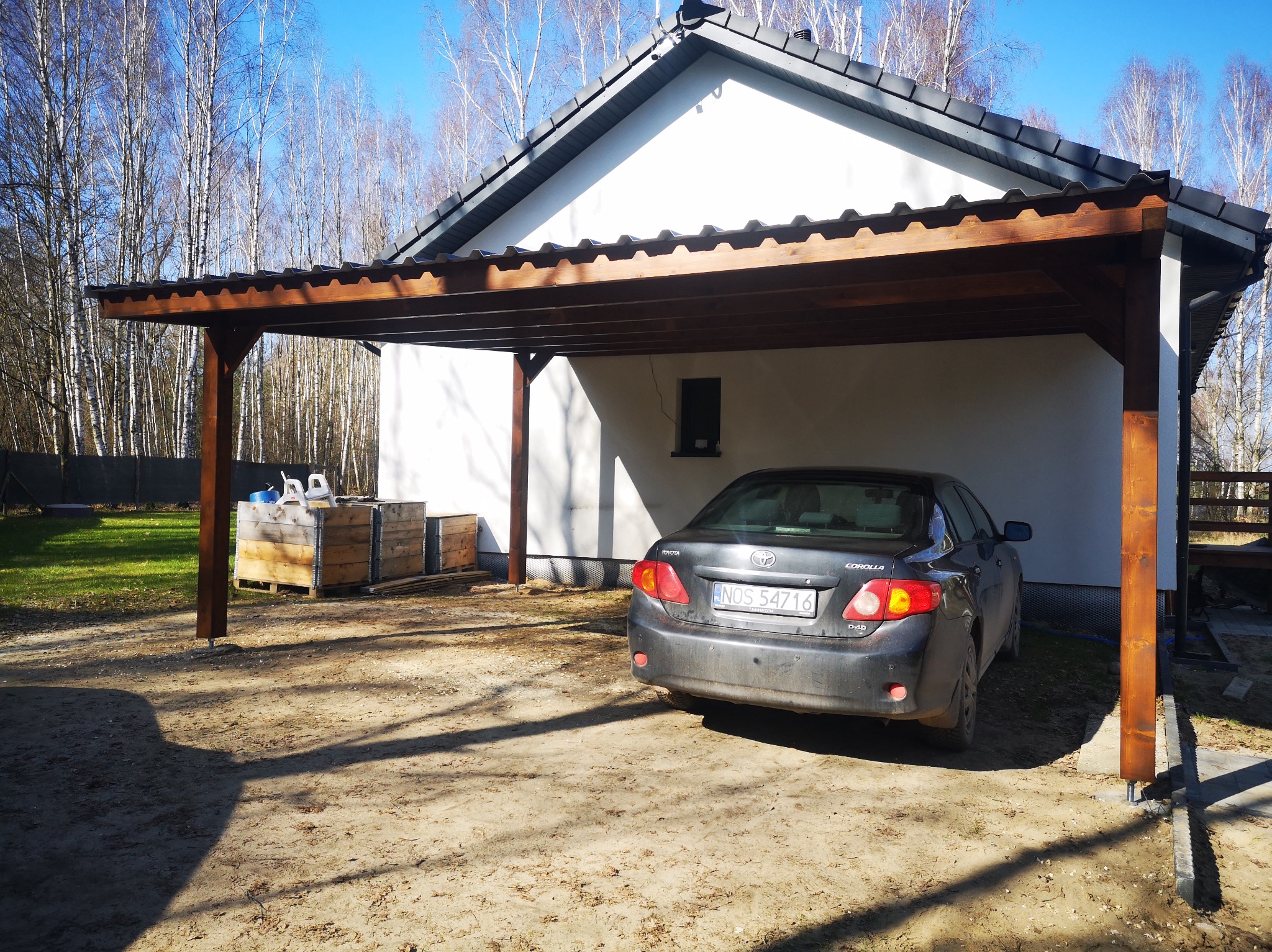 Drewniana wiata garażowa przylegająca do białego budynku, z zaparkowanym szarym samochodem. W tle drzewa bez liści i błękitne niebo. Widoczne skrzynie i cień.