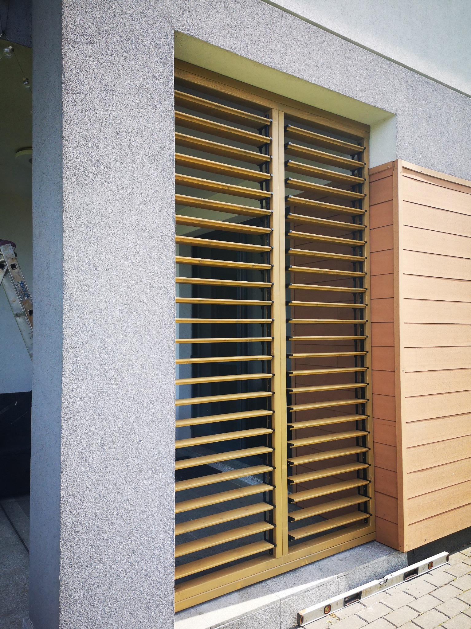 Nowoczesna zabudowa okienna z drewnianymi żaluzjami, widok z zewnątrz. Obok fragment elewacji z poziomym deskowaniem. Poziomica na posadzce.
