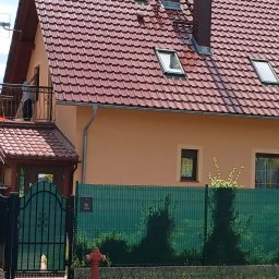 CleanRAT - Dom jednorodzinny po renowacji elewacji z nową, czerwoną dachówką. Widoczny fragment balkonu z osobą. Zieleń w tle i hydrant przed ogrodzeniem.