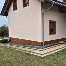 CleanRAT - Jasna elewacja domu jednorodzinnego z brązową podbitką i panelami słonecznymi na dachu, widok z boku na tle zielonego trawnika i kostki brukowej.