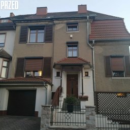 CleanRAT - Zdjęcie elewacji domu przed renowacją w Zgorzelcu. Widoczne zabrudzenia i ubytki tynku. Okna z brązowymi okiennicami. Dachówka w kolorze ceglastym.