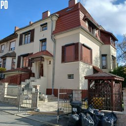 CleanRAT - Odnowiona, beżowa elewacja dwupiętrowego domu z brązowymi okiennicami i czerwoną dachówką po renowacji. Widoczne ślady prac porządkowych i remontowych.