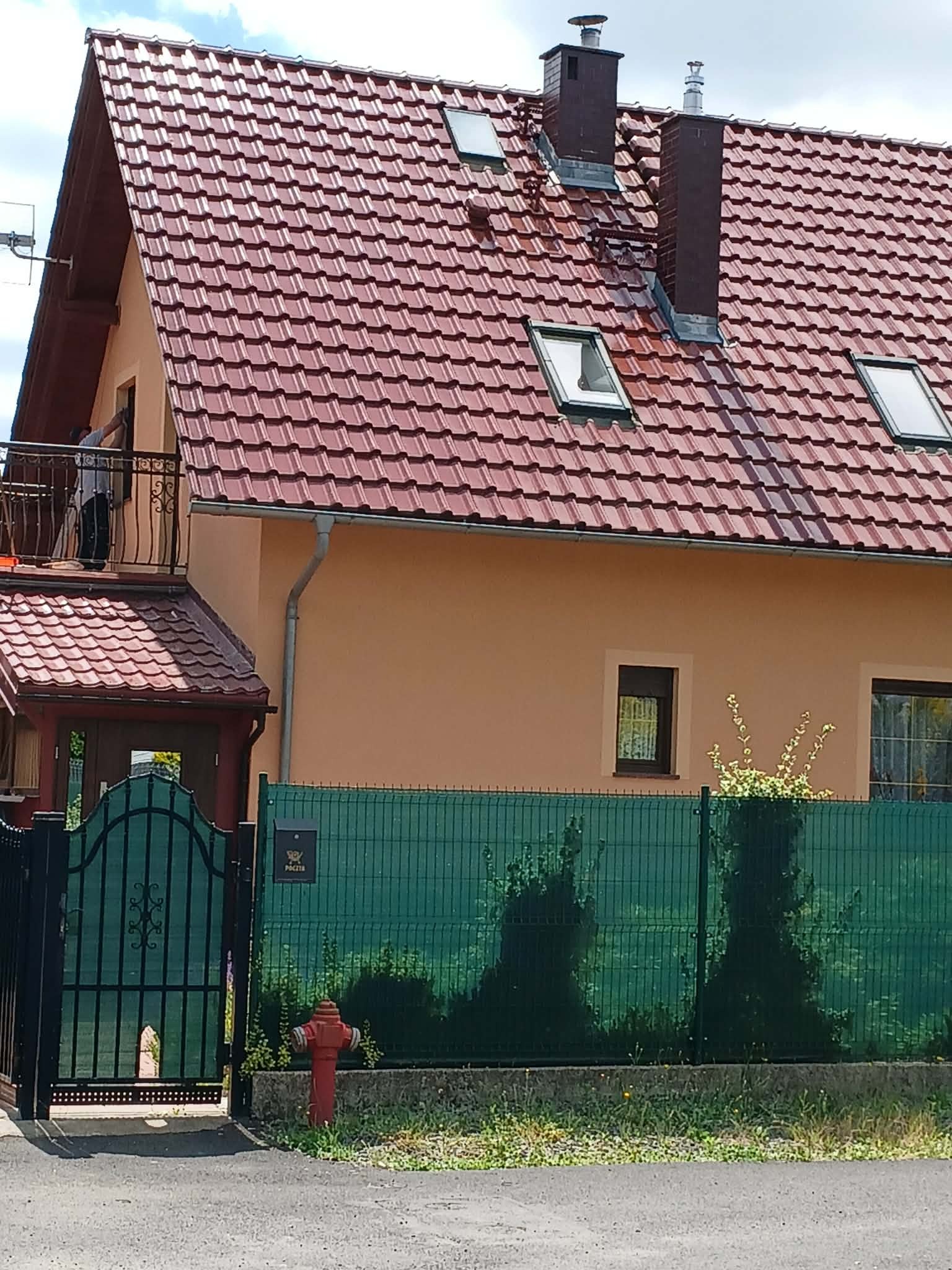 Dom jednorodzinny po renowacji elewacji z nową, czerwoną dachówką. Widoczny fragment balkonu z osobą. Zieleń w tle i hydrant przed ogrodzeniem.