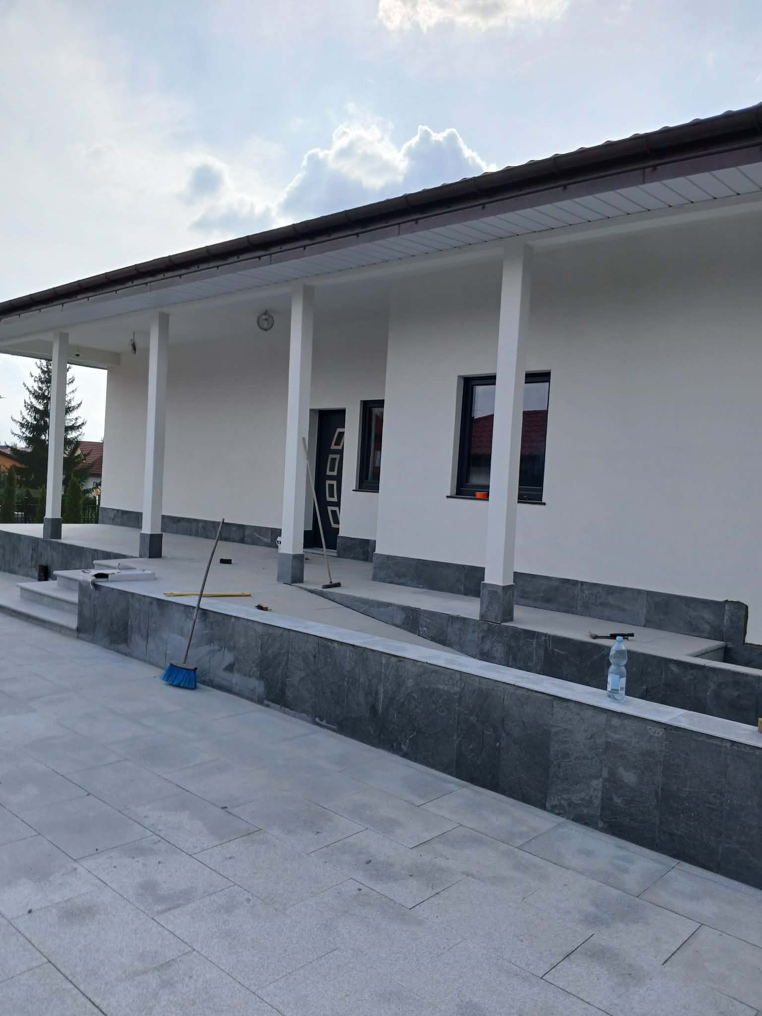 Biała elewacja domu po myciu i malowaniu, widoczne narzędzia pracy: miotła i szczotka. Szara podmurówka i chodnik. Błękitne niebo z chmurami w tle.