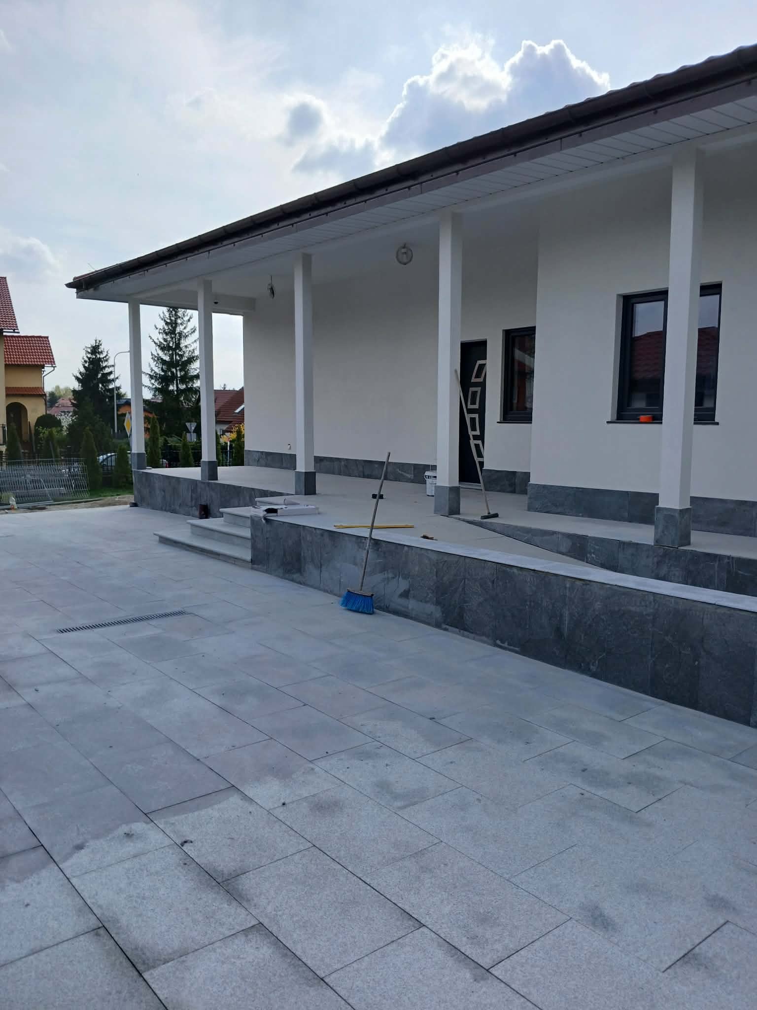 Biała elewacja domu po renowacji z szarym kamiennym wykończeniem. Widoczne narzędzia: drabina, szczotka. Ujęcie z poziomu chodnika z szarej kostki.