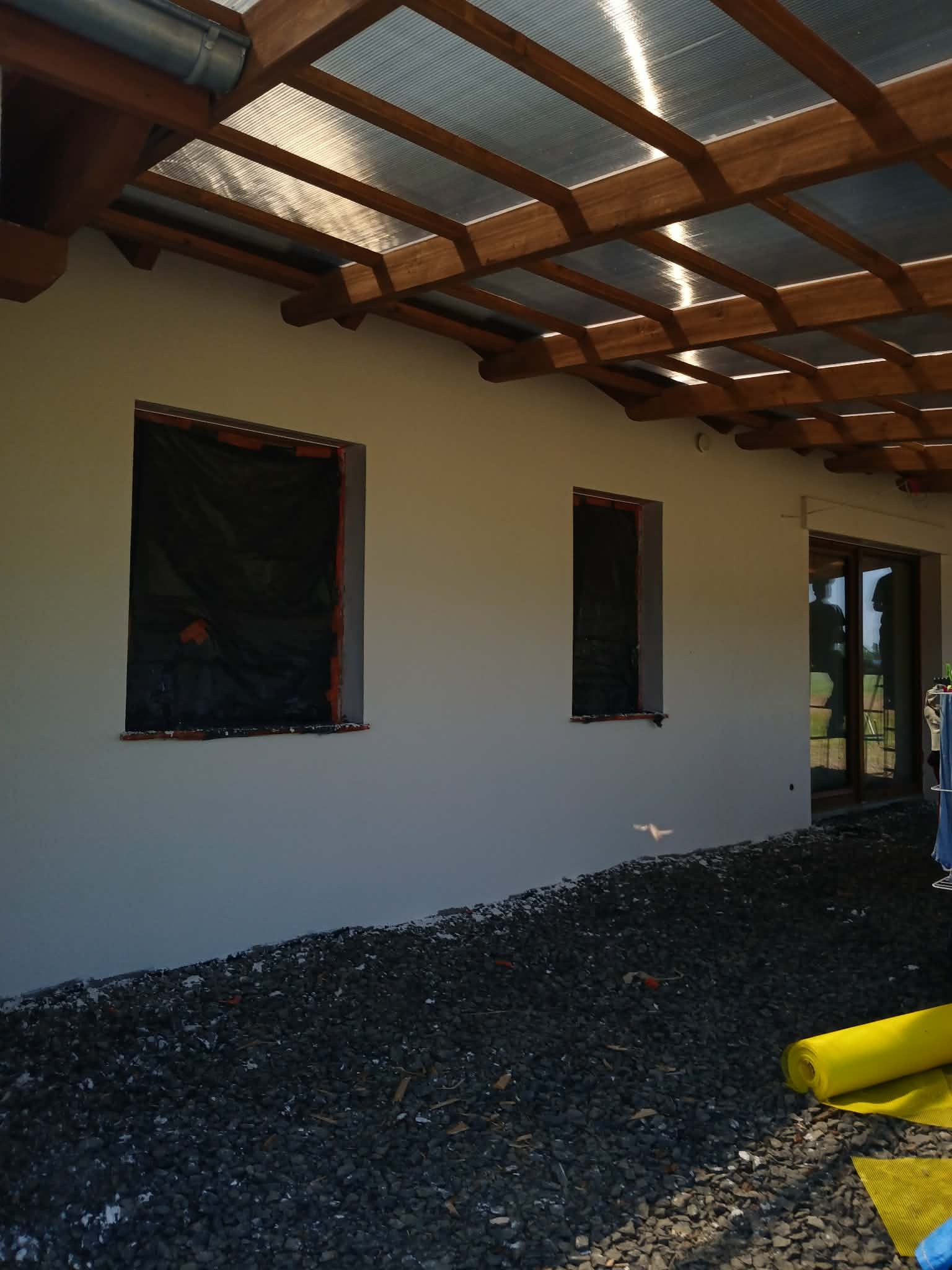 Świeżo otynkowana elewacja domu jednorodzinnego z zabezpieczonymi oknami folią. Widoczne kamienie i żółta siatka na ziemi. Zadaszenie z drewnianych belek.