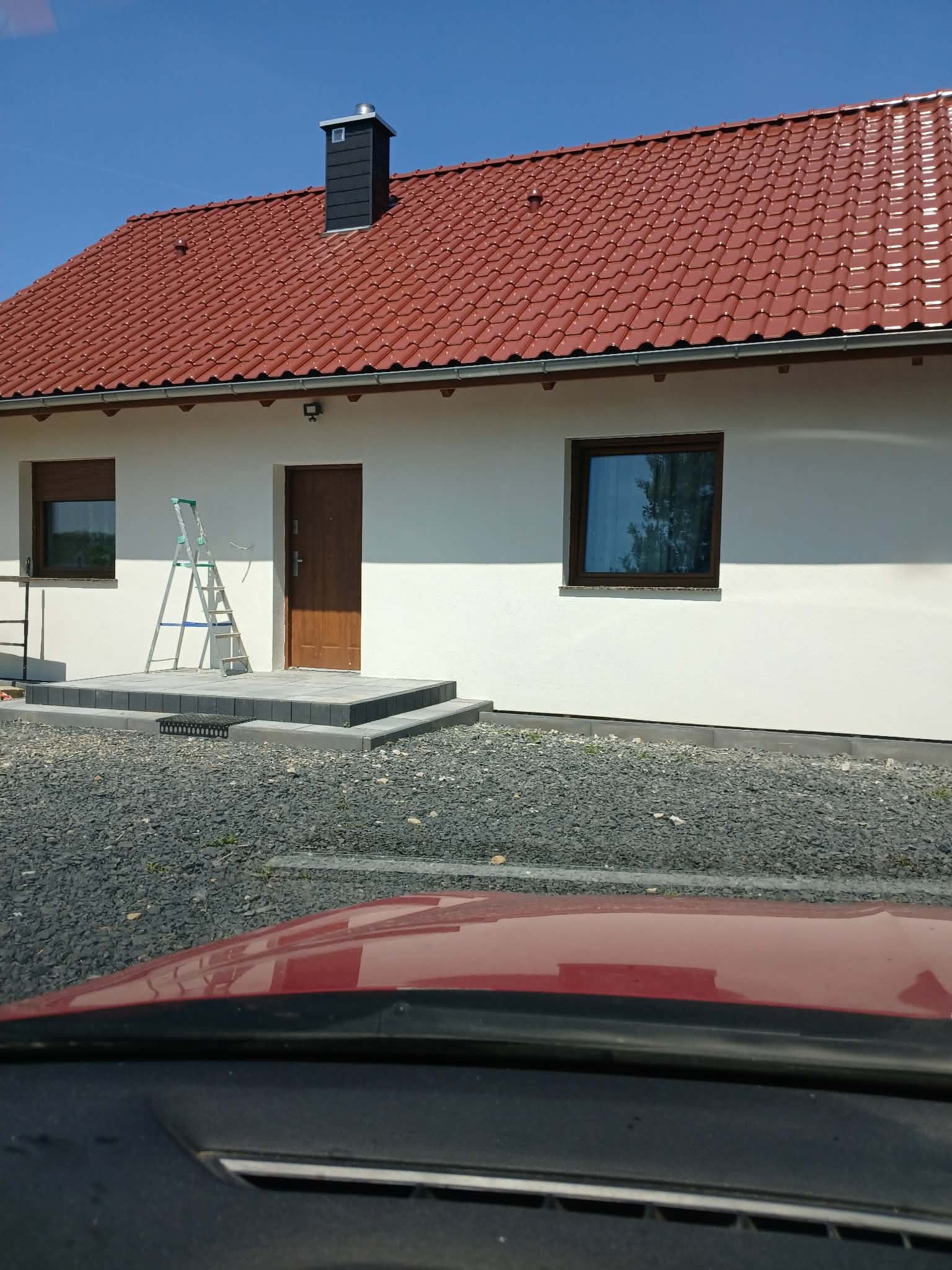 Dom jednorodzinny po tynkowaniu elewacji w Mierzwinie. Widoczna drabina oparta o ścianę, brązowe okna i drzwi. Czerwona dachówka i czarny komin.