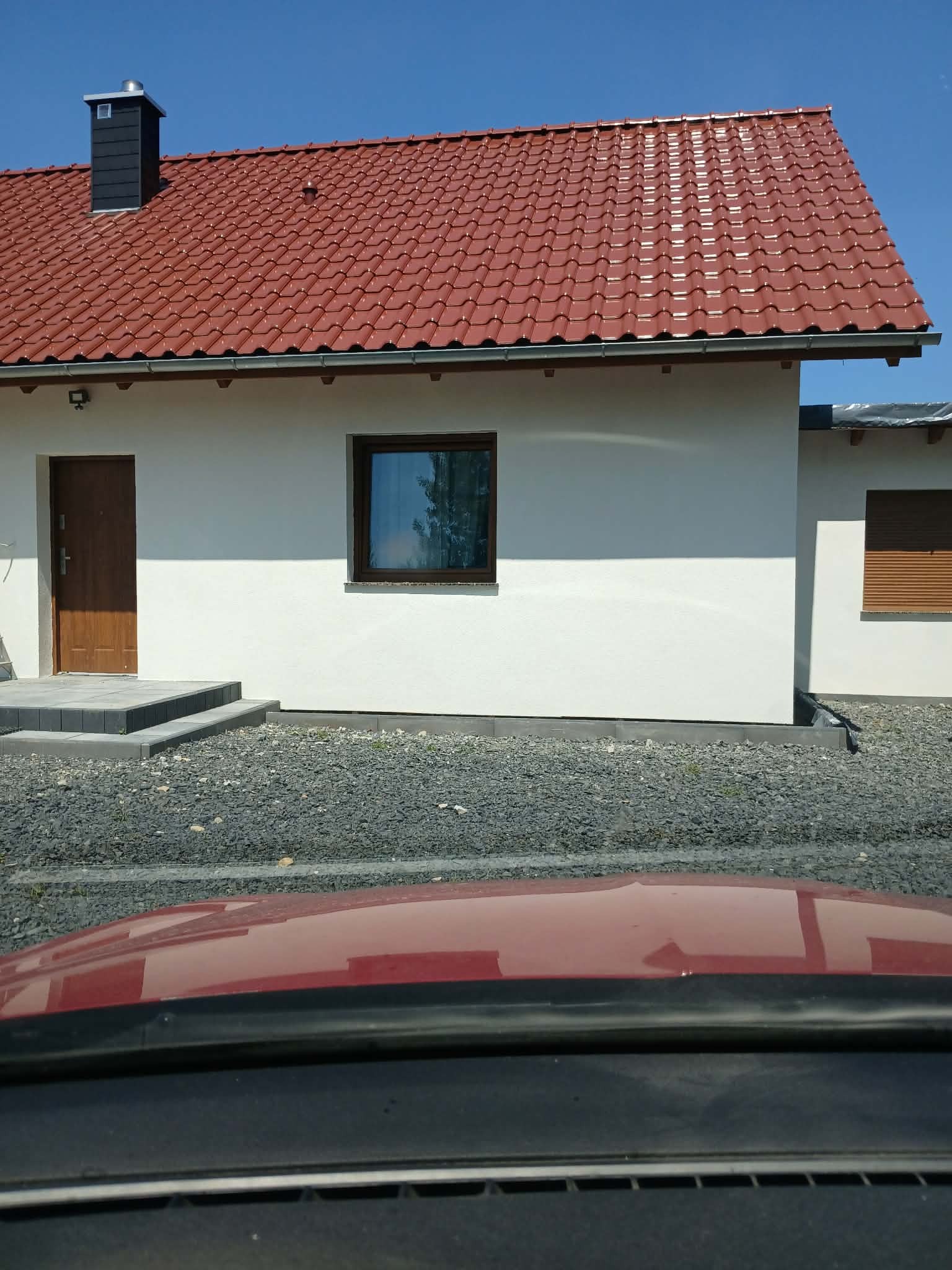 Jasny dom jednorodzinny z czerwoną dachówką i ciemnymi oknami, świeżo po tynkowaniu, widok zza szyby samochodu, z kamienistym podłożem.