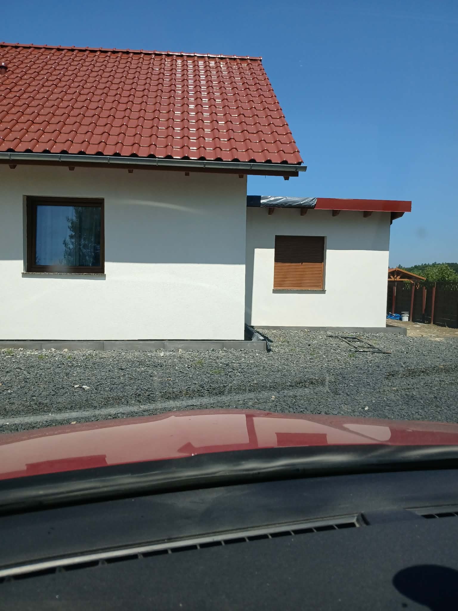 Biała elewacja domu jednorodzinnego z brązowymi oknami i dachem z czerwonej dachówki, widok zza szyby samochodu, kamieniste podłoże.