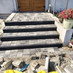 U Szymona - Ułożone schody wejściowe z kostki brukowej w odcieniach szarości i czerni, z kwiatami w donicy po prawej stronie. Widoczne narzędzia i materiały budowlane.