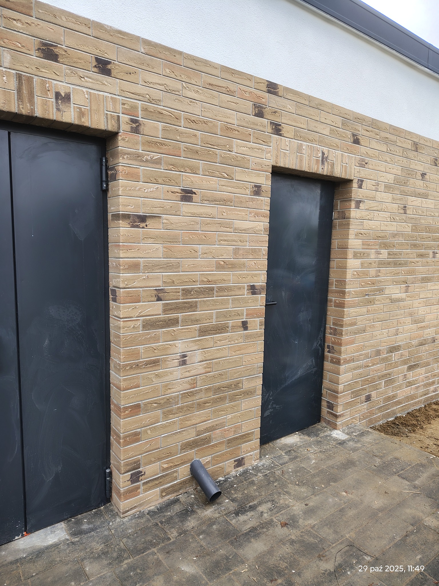 Elewacja budynku z cegły klinkierowej w odcieniach beżu i brązu, z dwoma ciemnymi, metalowymi drzwiami. Widoczny fragment chodnika z kostki brukowej i rura spustowa.