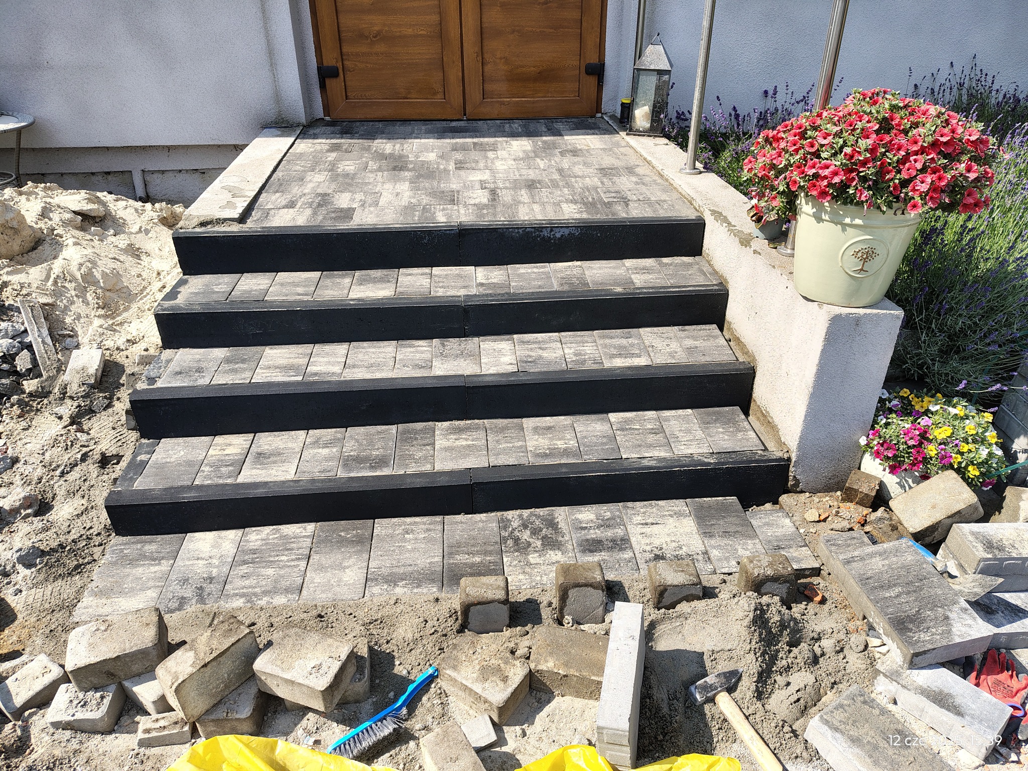 Ułożone schody wejściowe z kostki brukowej w odcieniach szarości i czerni, z kwiatami w donicy po prawej stronie. Widoczne narzędzia i materiały budowlane.