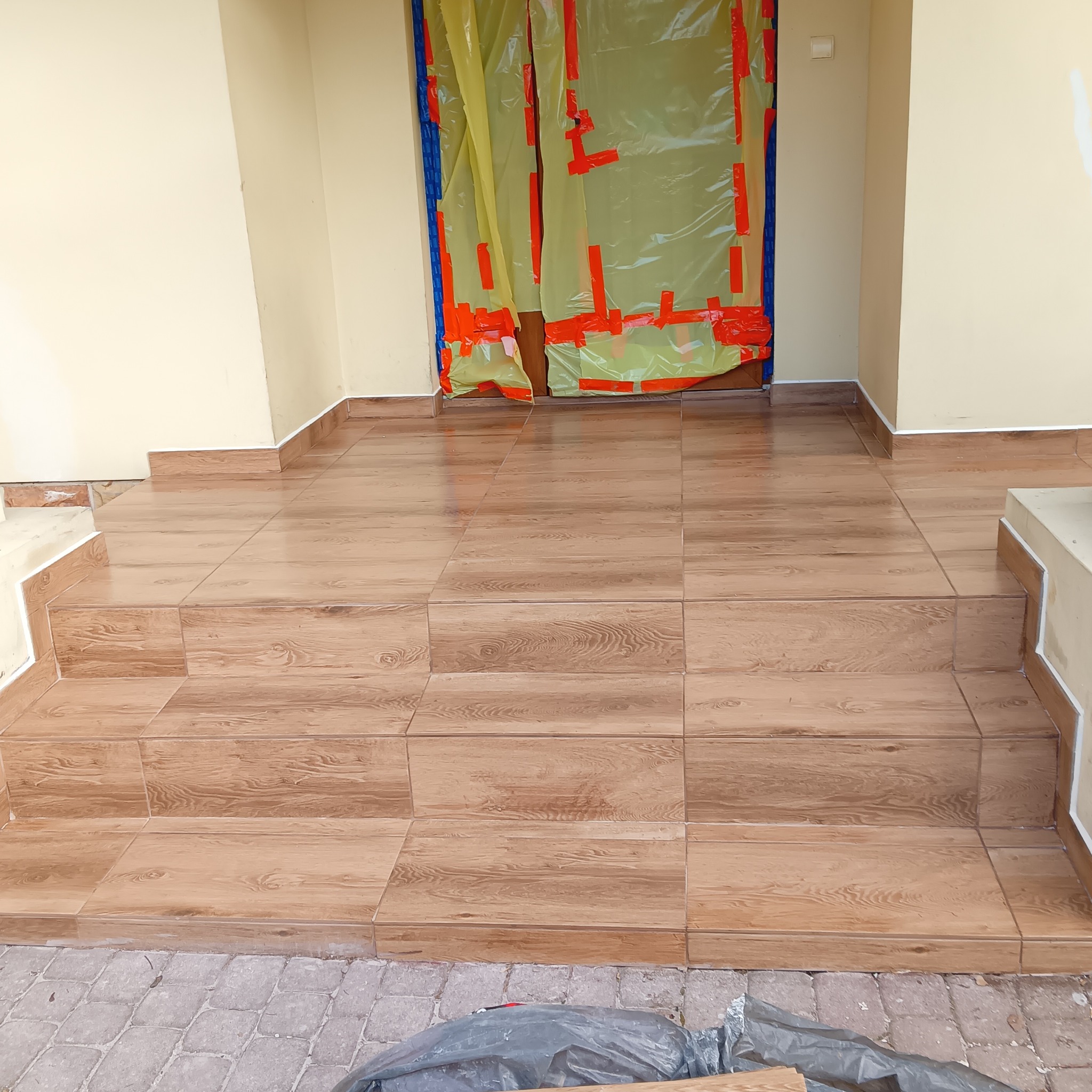 Wykończone płytkami schody wejściowe do domu, z zabezpieczonymi folią drzwiami. Płytki imitują drewno, jasne ściany. Widoczne detale wykończenia i zabezpieczenia.