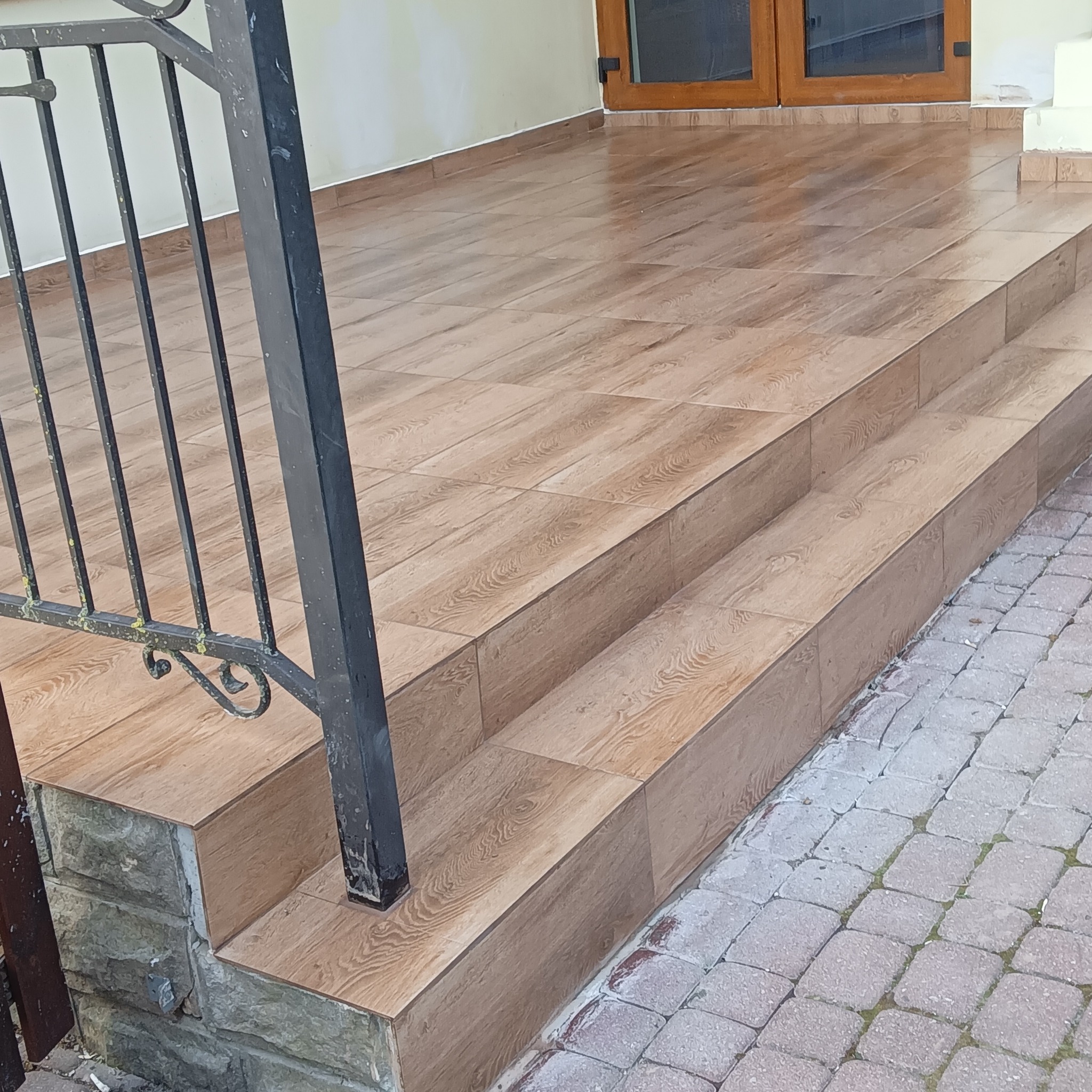 Wykończone płytkami schody wejściowe z metalową barierką. Płytki imitują drewno, a podłoże wyłożone jest kostką brukową. Widoczne detale wykończenia.