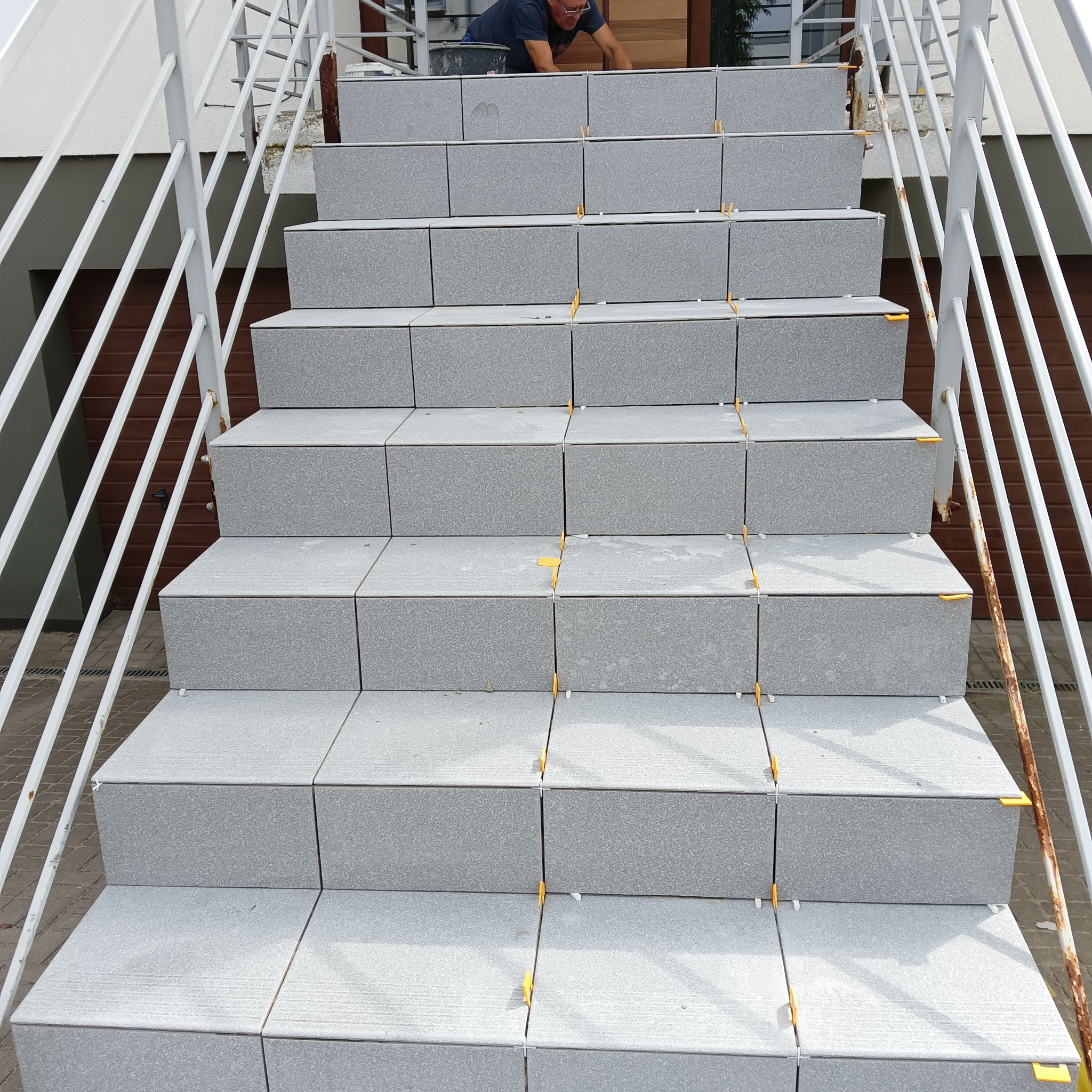 Wykończone płytkami schody zewnętrzne z białymi barierkami, widoczne żółte krzyżyki dystansowe. Glazurnik w tle. Szare płytki na schodach.