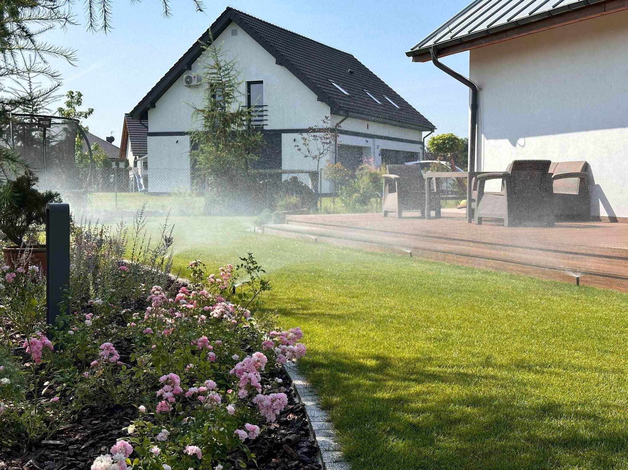 Ogród z automatycznym nawadnianiem w słoneczny dzień. Widoczny trawnik, rabaty z różowymi kwiatami, drewniany taras i dom w tle. Woda zraszająca trawnik.