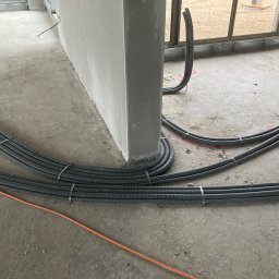 Konrad Hartmann - Szare rury instalacji elektrycznej ułożone na surowym betonie wzdłuż ściany, zabezpieczone metalowymi opaskami. Widoczne okno w tle i pomarańczowy kabel.