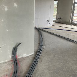 Konrad Hartmann - Szare rury instalacji elektrycznej ułożone wzdłuż ściany w surowym wnętrzu budynku w trakcie budowy. Widoczne kable i puszki elektryczne.