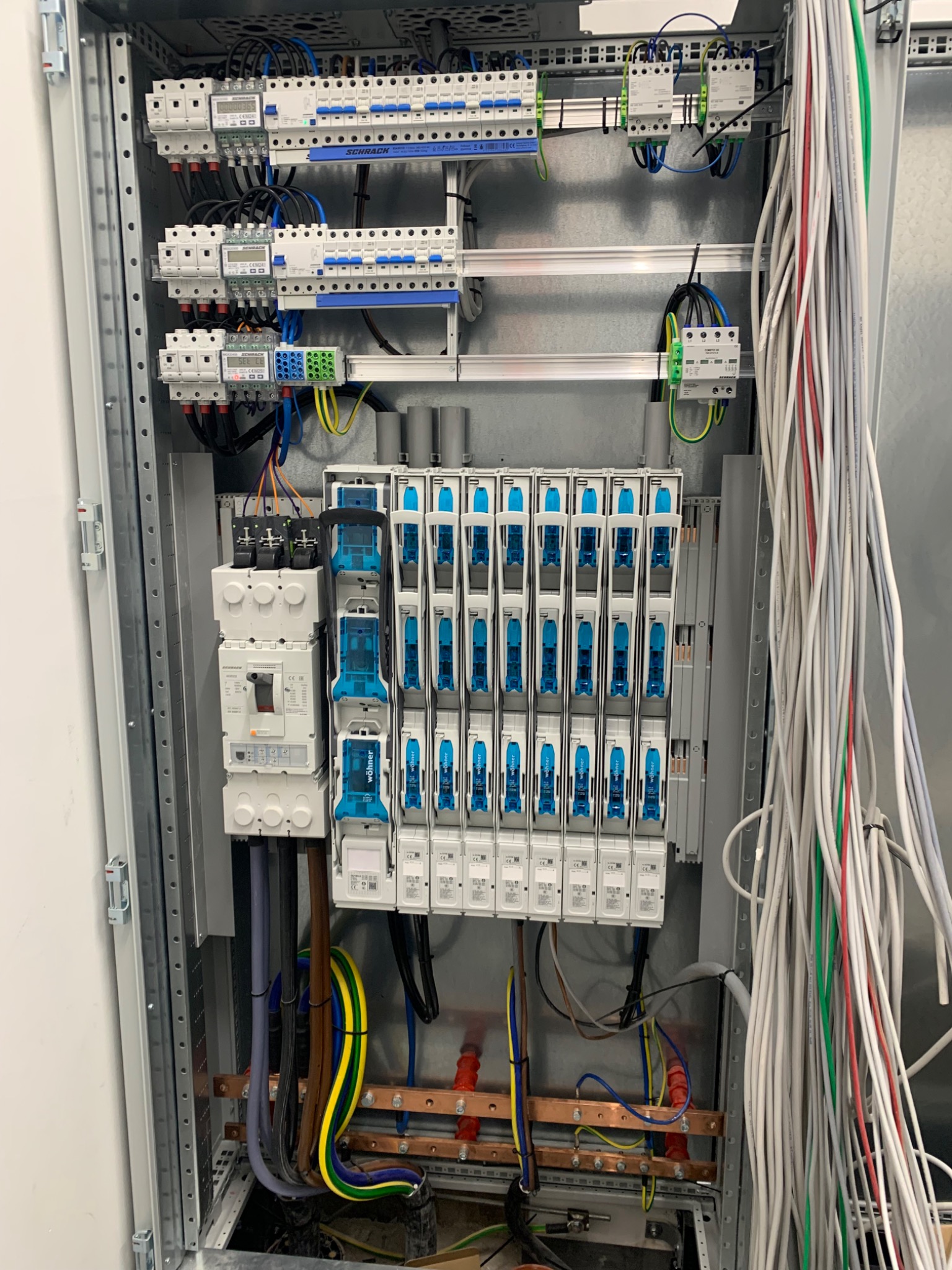 Rozdzielnia elektryczna z zainstalowanymi bezpiecznikami, licznikami i okablowaniem. Widoczne elementy montażowe i przewody w różnych kolorach.