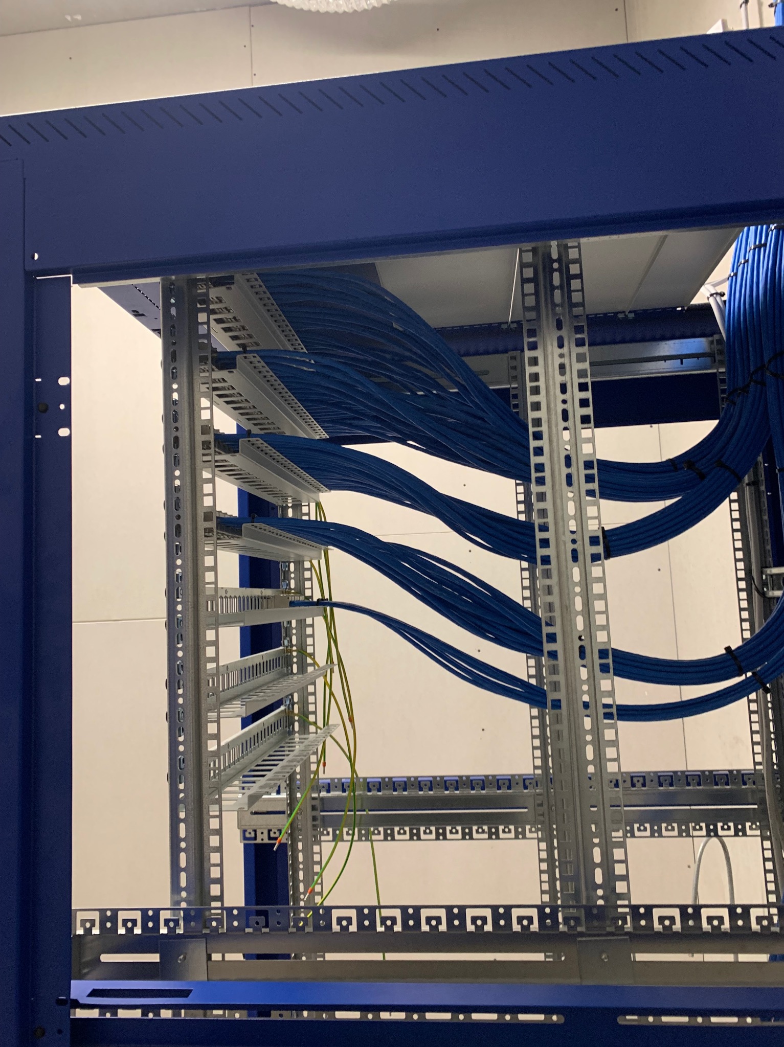 Niebieska szafa rack z uporządkowanymi kablami sieciowymi w kolorze niebieskim i zielonym. Widoczne metalowe elementy konstrukcyjne i białe tło.