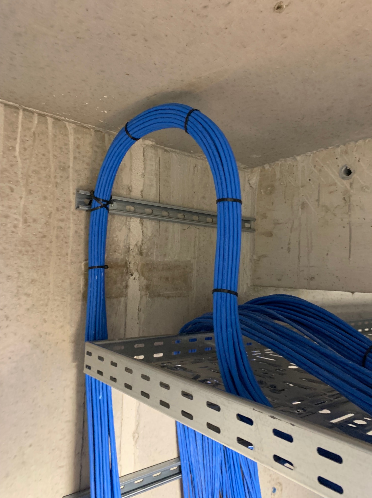 Niebieskie przewody elektryczne ułożone w łuk na metalowej drabince, przymocowane do surowej, betonowej ściany. Widoczne czarne opaski zaciskowe.