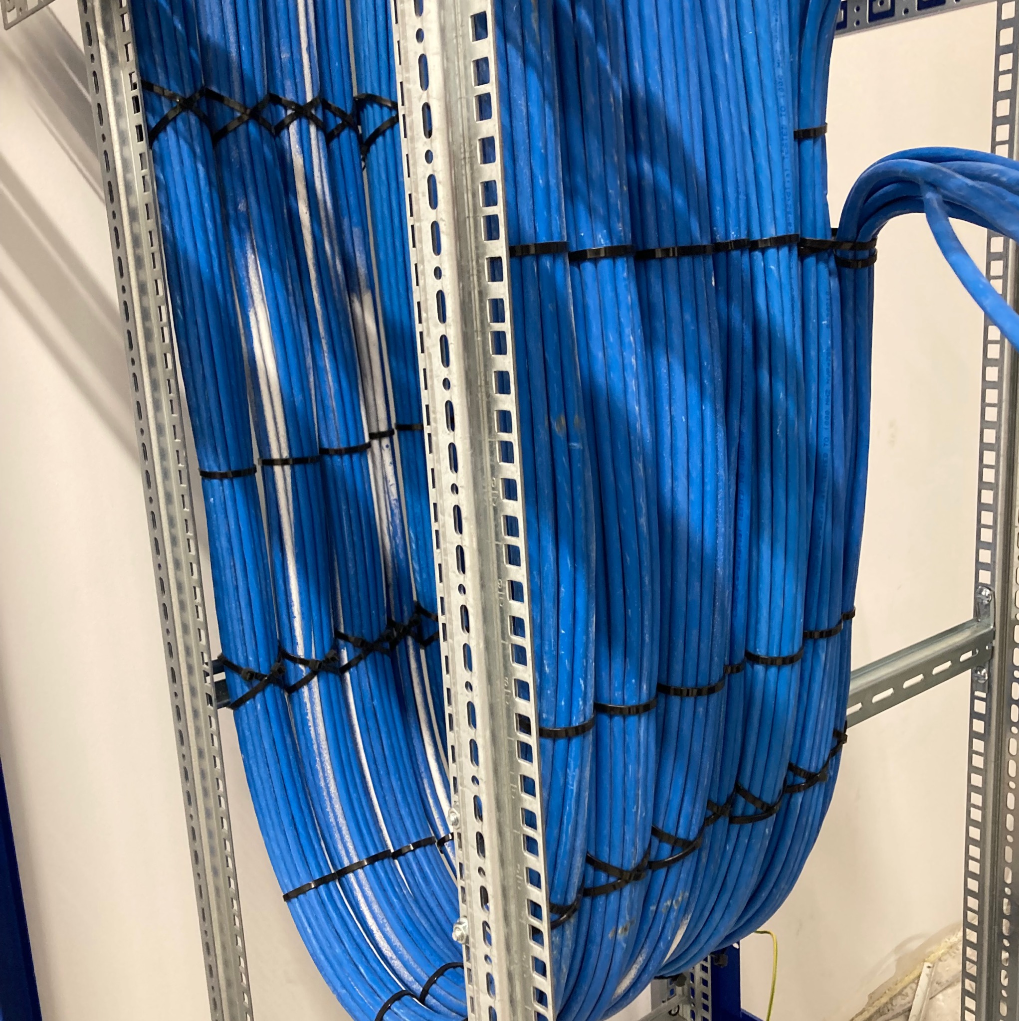 Równolegle ułożone niebieskie przewody w metalowej konstrukcji, zabezpieczone czarnymi opaskami zaciskowymi. Widoczne elementy mocujące do ściany.