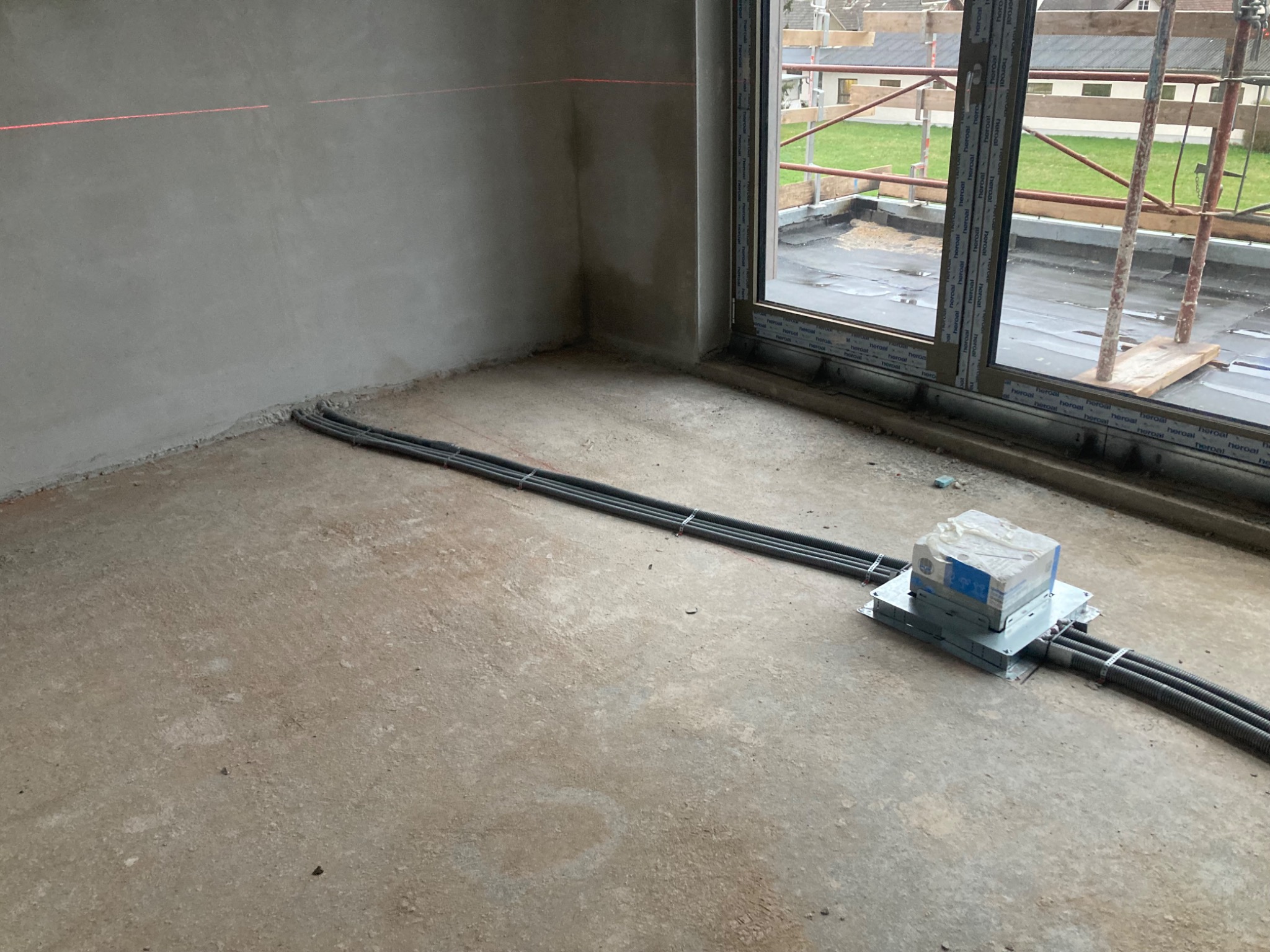 Wnętrze w trakcie prac wykończeniowych: widoczne kable elektryczne w peszlach ułożone na podłodze, skrzynka elektryczna i poziomica laserowa na ścianie. Okno z widokiem na zewnątrz.