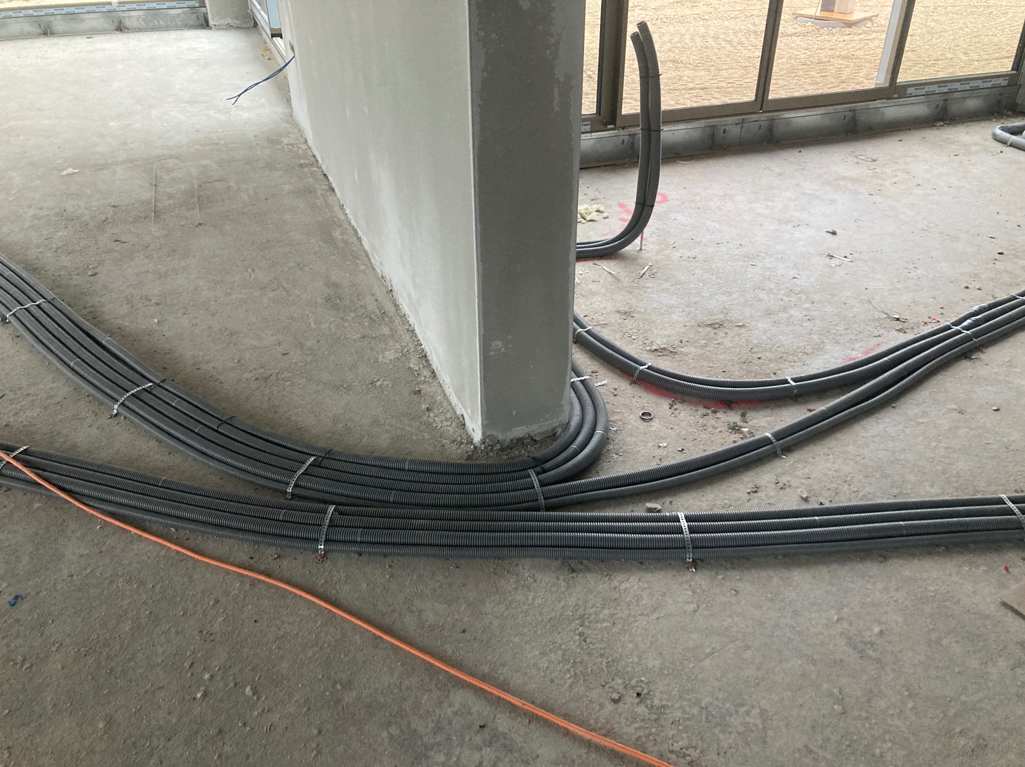 Szare rury instalacji elektrycznej ułożone na surowym betonie wzdłuż ściany, zabezpieczone metalowymi opaskami. Widoczne okno w tle i pomarańczowy kabel.