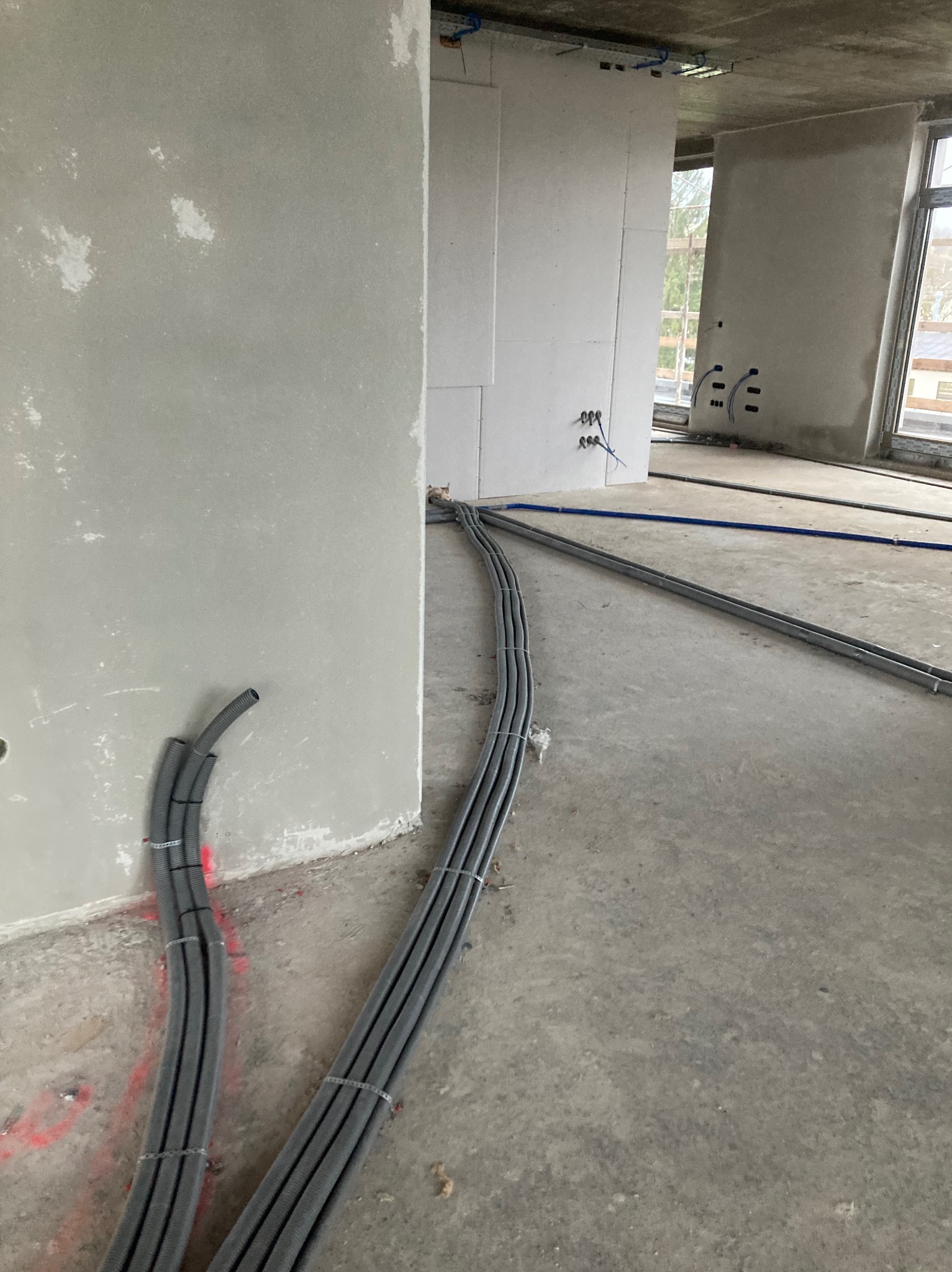 Szare rury instalacji elektrycznej ułożone wzdłuż ściany w surowym wnętrzu budynku w trakcie budowy. Widoczne kable i puszki elektryczne.