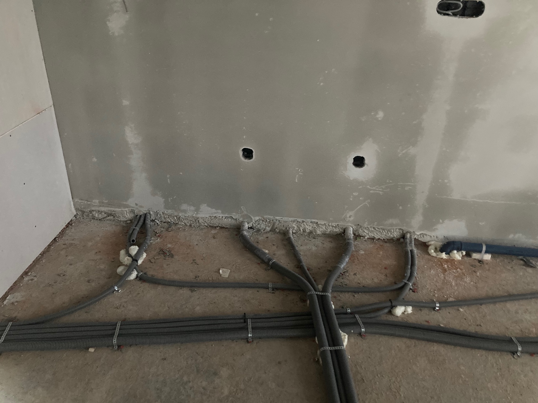 Szare, karbowane rury instalacji elektrycznej ułożone na surowym podłożu, prowadzące do otworów w ścianie pokrytej gładzią. Widoczne uchwyty mocujące i pianka montażowa.