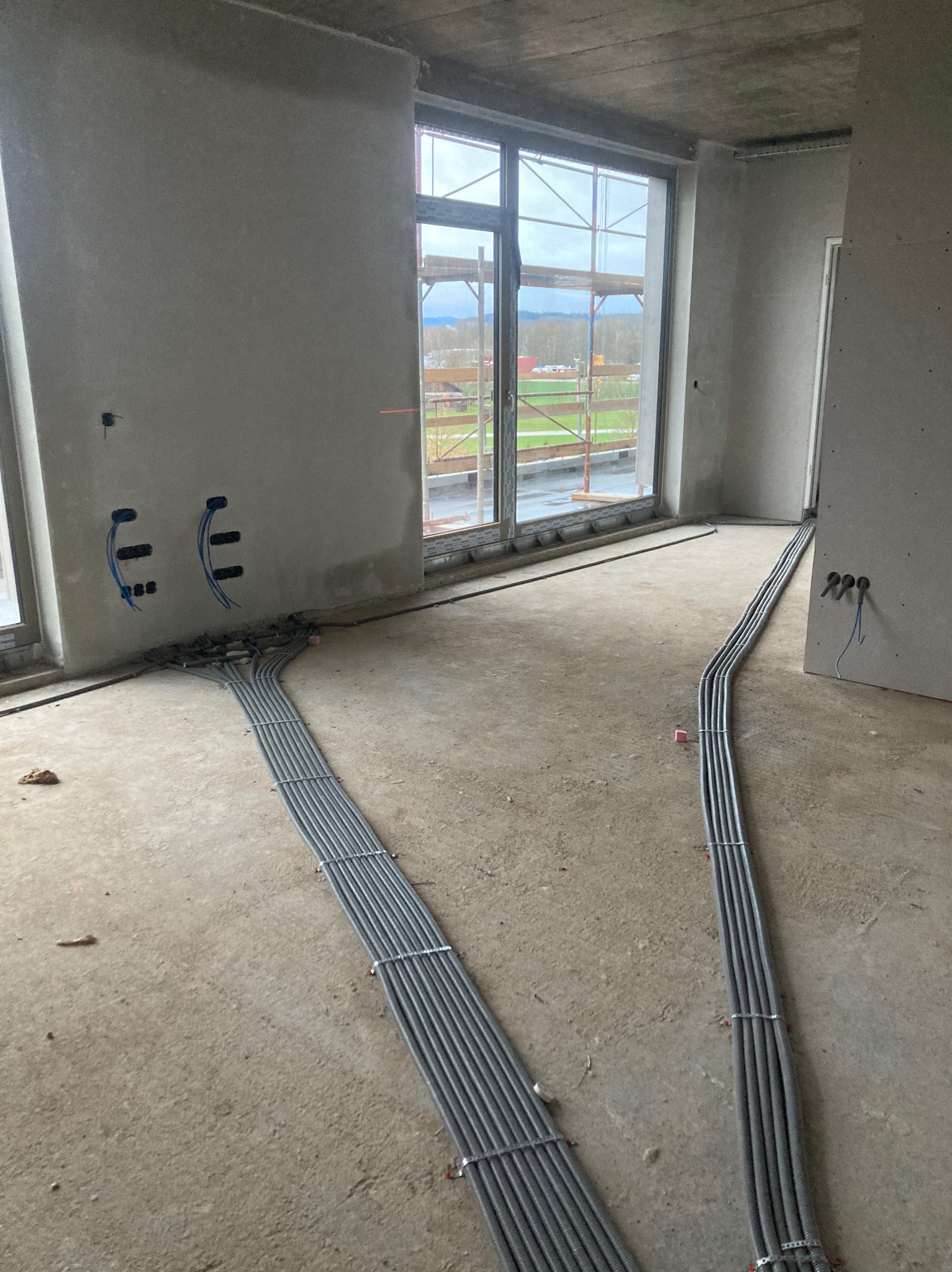Szare rury instalacji elektrycznej ułożone równolegle na surowym betonie w nowym budynku. Widoczne otwory w ścianach i okno z widokiem na zewnątrz.
