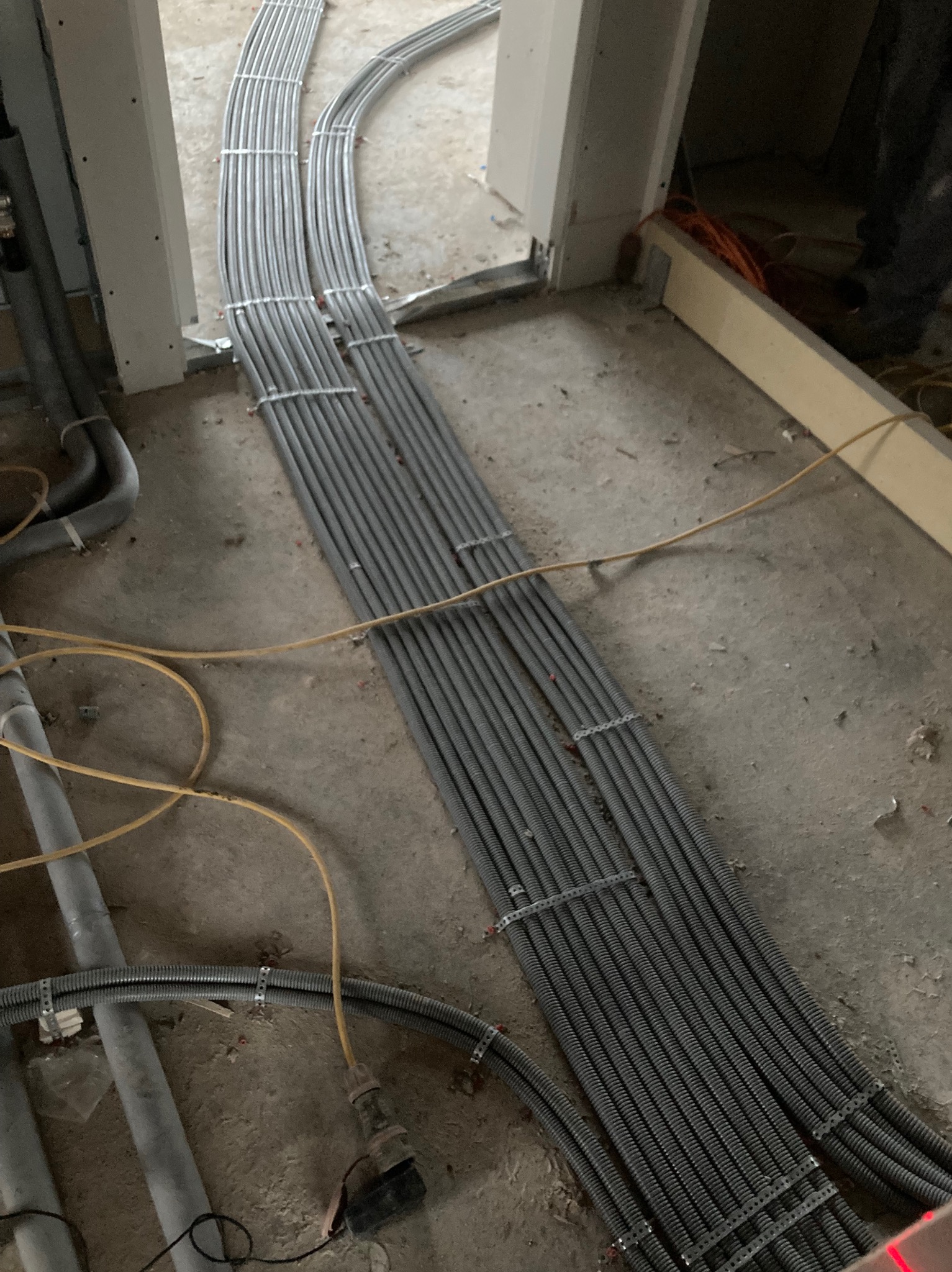 Szereg szarych, elastycznych rur instalacyjnych ułożonych równolegle na surowym podłożu w trakcie prac wykończeniowych, zabezpieczonych metalowymi obejmami.