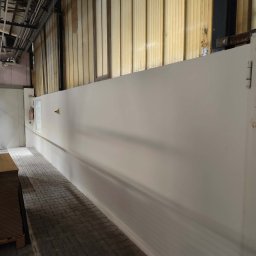 Mariola laskowska - Świeżo pomalowana, długa ściana w hali przemysłowej, widoczna z perspektywy podłogi z kostki brukowej. W tle instalacje pod sufitem i paleta z płytami.