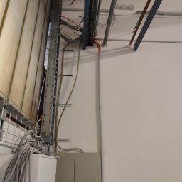 Mariola laskowska - Instalacja elektryczna w budynku: kable w korytkach, puszki elektryczne na ścianie, widoczne rury pod sufitem. Prace instalacyjne w surowym wnętrzu.