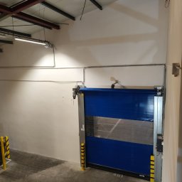 Mariola laskowska - Błękitna brama przemysłowa z żółto-czarnymi odbojnikami w hali magazynowej. Widoczne instalacje pod sufitem i betonowa posadzka. Ściany pomalowane na jasny kolor.