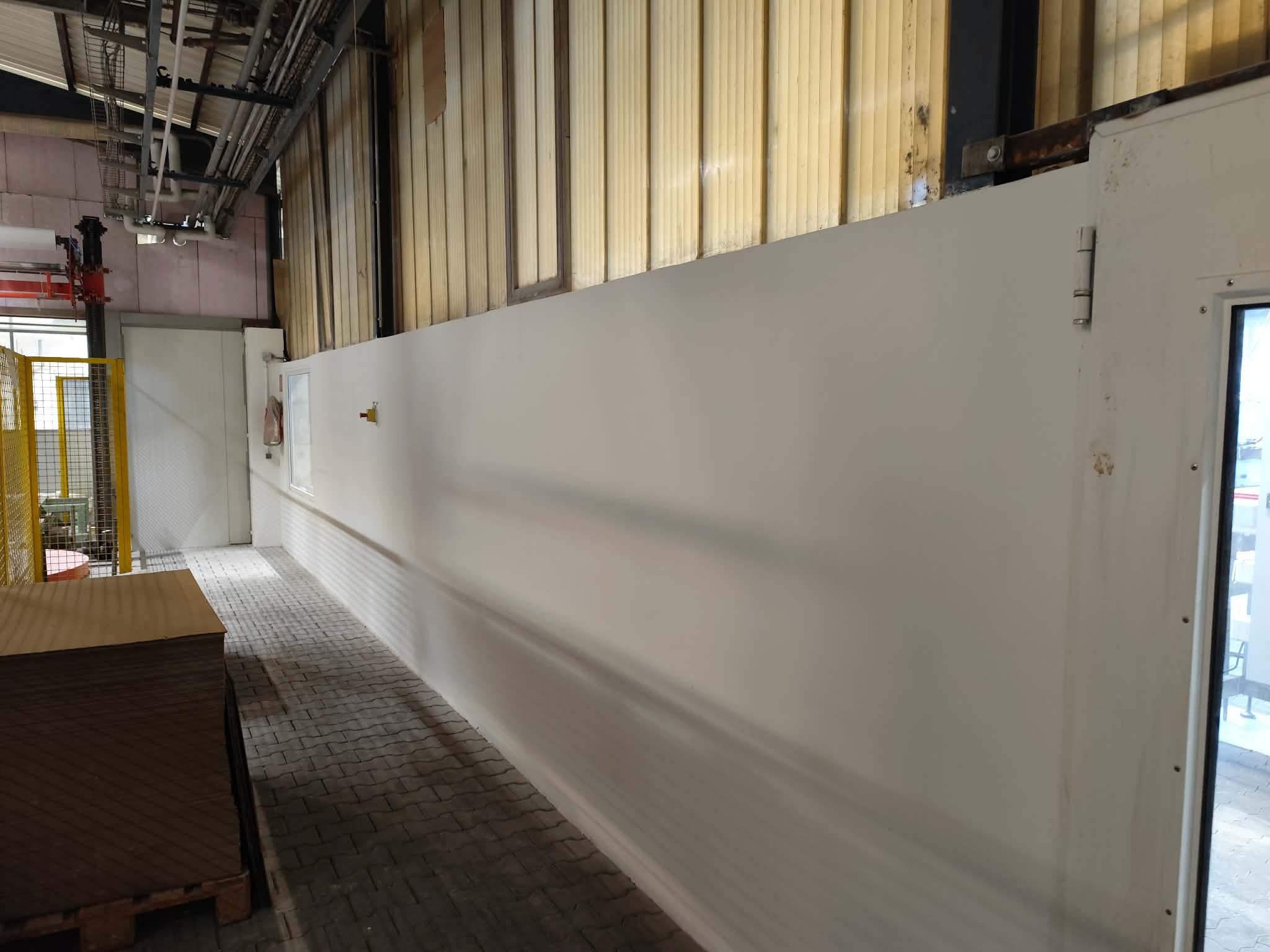Świeżo pomalowana, długa ściana w hali przemysłowej, widoczna z perspektywy podłogi z kostki brukowej. W tle instalacje pod sufitem i paleta z płytami.