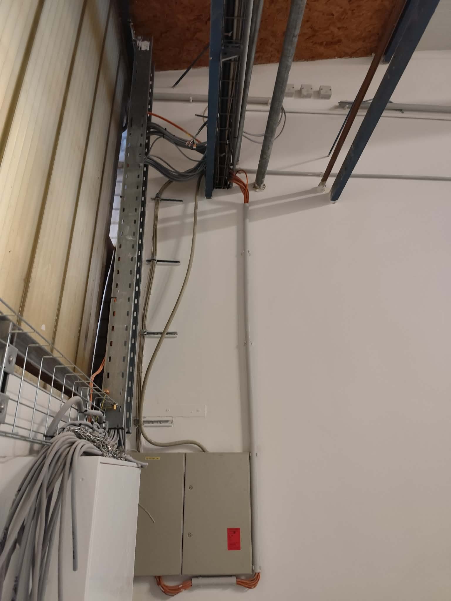 Instalacja elektryczna w budynku: kable w korytkach, puszki elektryczne na ścianie, widoczne rury pod sufitem. Prace instalacyjne w surowym wnętrzu.