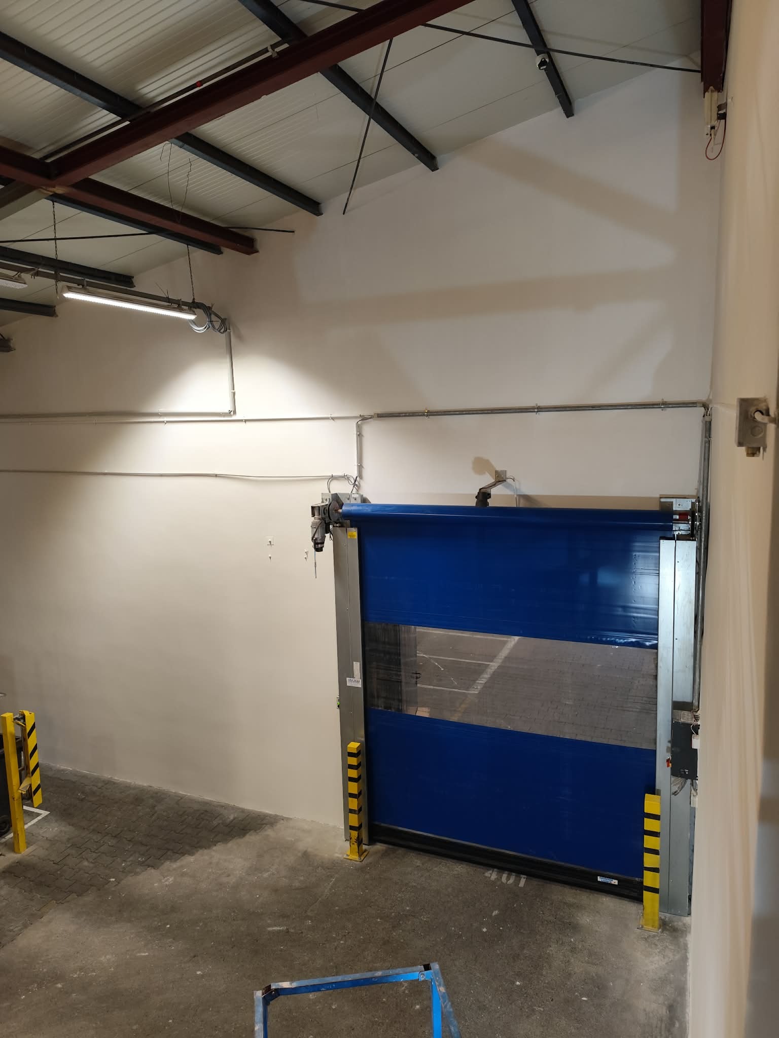 Błękitna brama przemysłowa z żółto-czarnymi odbojnikami w hali magazynowej. Widoczne instalacje pod sufitem i betonowa posadzka. Ściany pomalowane na jasny kolor.