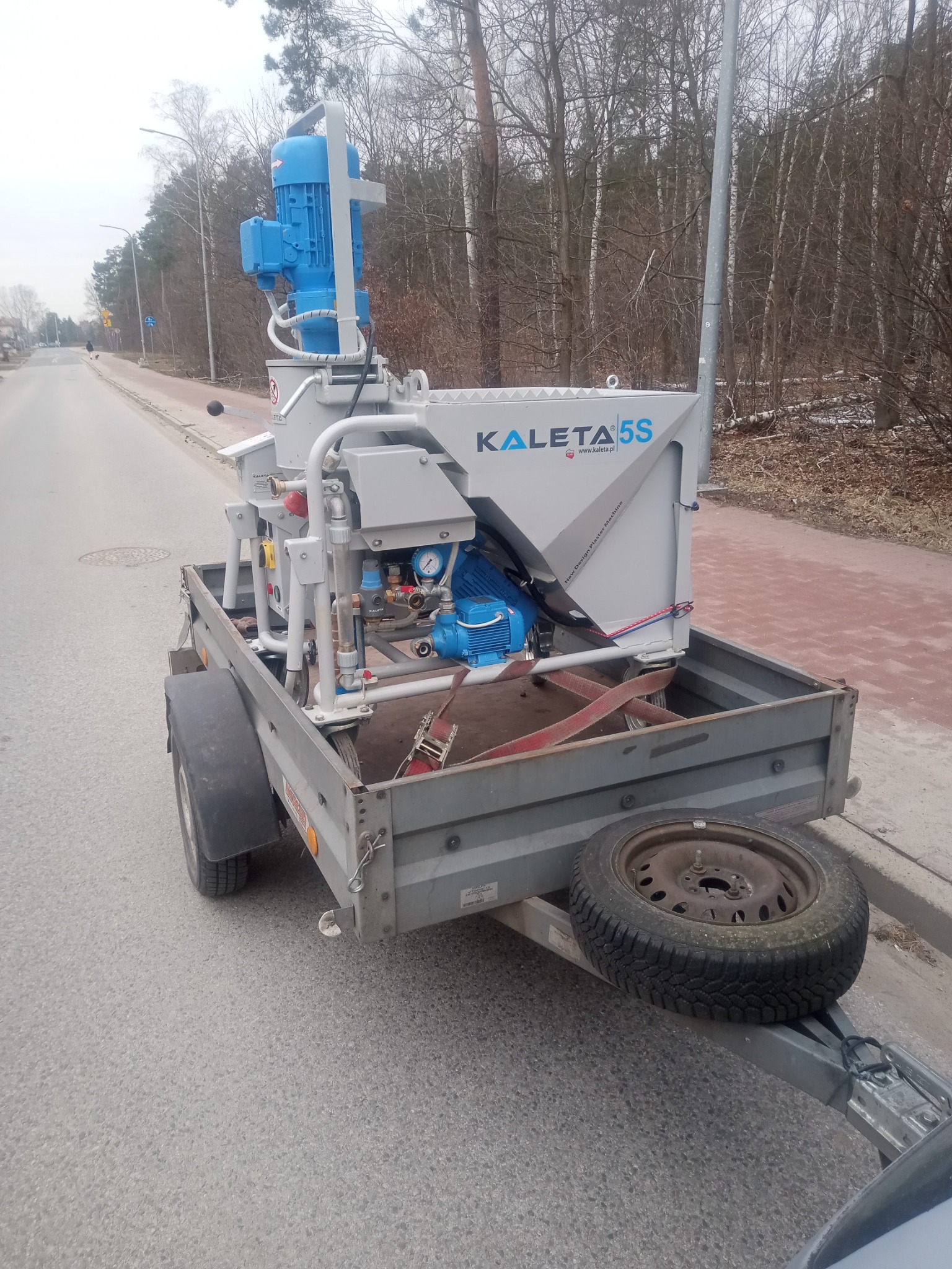 Maszyna do tynkowania Kaleta 5S na przyczepie, widok z boku na tle drogi i drzew. Urządzenie w kolorach szarym i niebieskim, z widocznymi elementami hydraulicznymi.