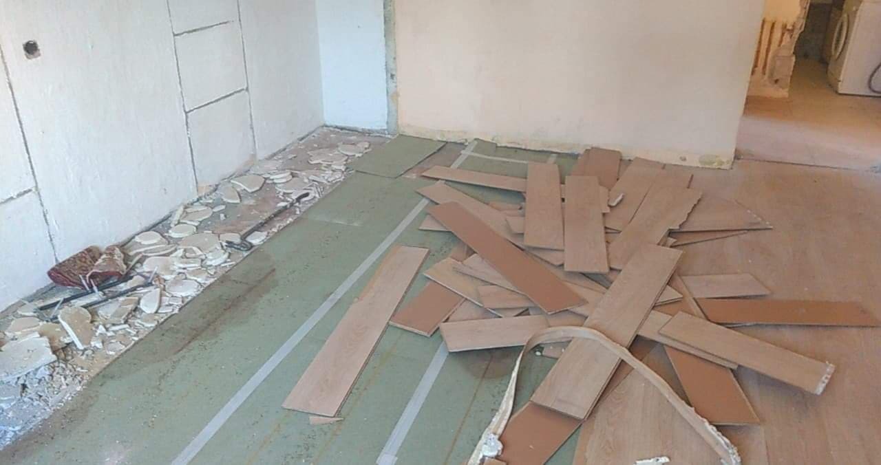 Demontaż podłogi: fragmenty paneli i gruzu na podłożu. Widoczne ślady po starej podłodze i przygotowanie do nowej. Remont w trakcie, wnętrze mieszkania.