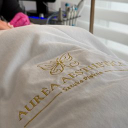 Aurea Aesthetica Weronika Szkolak - Koc z haftowanym logo salonu kosmetycznego Aurea Aesthetica, w tle monitor z logo i narzędzia kosmetyczne. Ujęcie z perspektywy klienta leżącego na leżance.