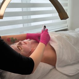 Aurea Aesthetica Weronika Szkolak - Klientka z maską na twarzy leży na łóżku kosmetycznym, podczas gdy kosmetyczka w różowych rękawiczkach wykonuje zabieg. Widoczna lampa kosmetyczna i tatuaż na ramieniu.