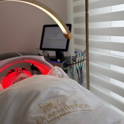 Aurea Aesthetica Weronika Szkolak - Klientka poddaje się zabiegowi na ciało w salonie kosmetycznym, oświetlona lampą LED. Widoczne narzędzia i monitor z logo salonu Aurea Aesthetica.