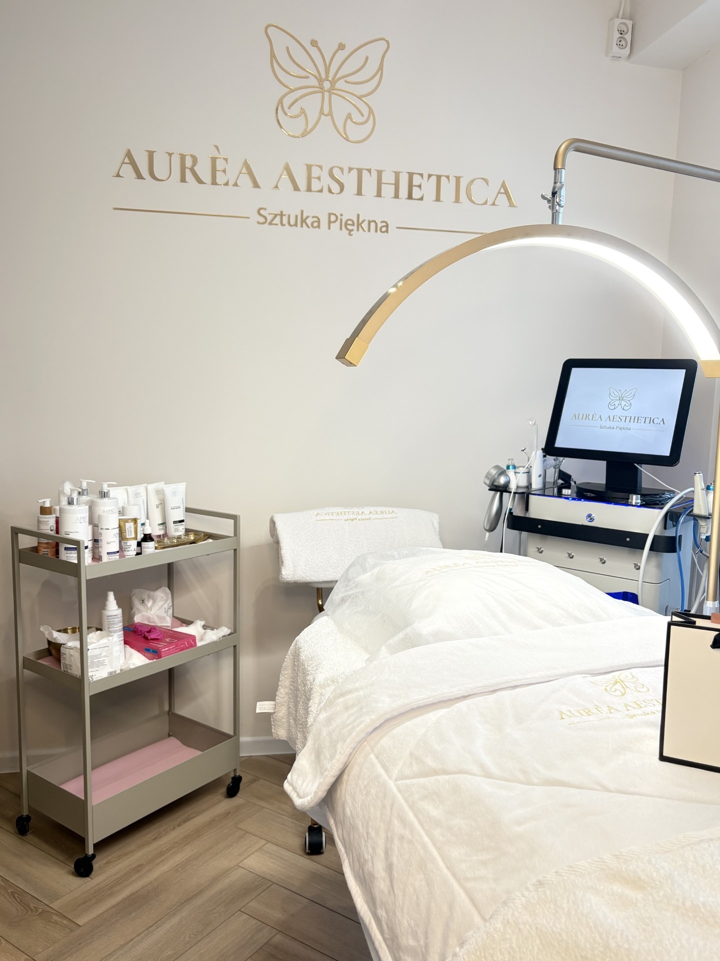 Jasne wnętrze salonu z leżanką, wózkiem z kosmetykami i aparaturą. Logo firmy w złotym kolorze na ścianie i monitorze. Minimalistyczny wystrój.