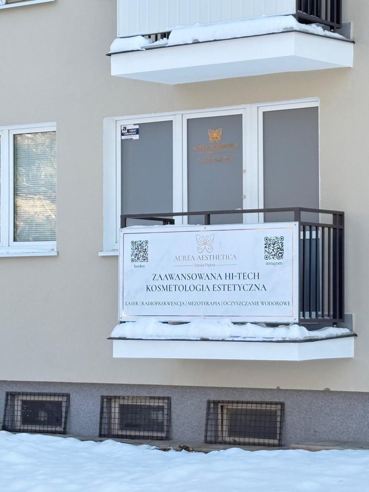 Zewnętrzny widok salonu kosmetycznego Aurea Aesthetica z balkonem, na którym umieszczono baner reklamowy z ofertą zabiegów hi-tech. Zima, śnieg na balkonie.