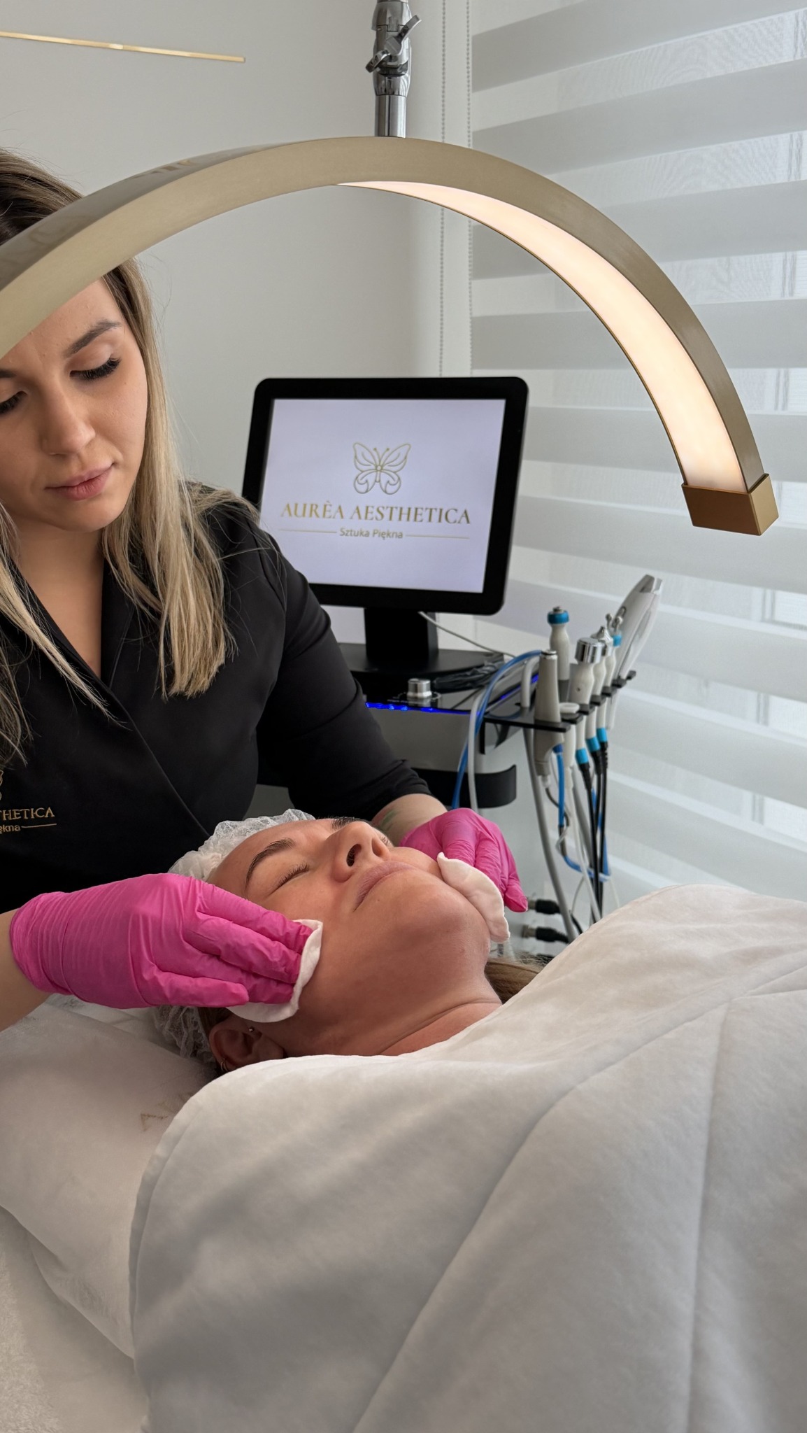 Klientka w trakcie zabiegu oczyszczania twarzy w salonie kosmetycznym. Kosmetolog w różowych rękawiczkach delikatnie oczyszcza skórę w jasnym, nowoczesnym wnętrzu.