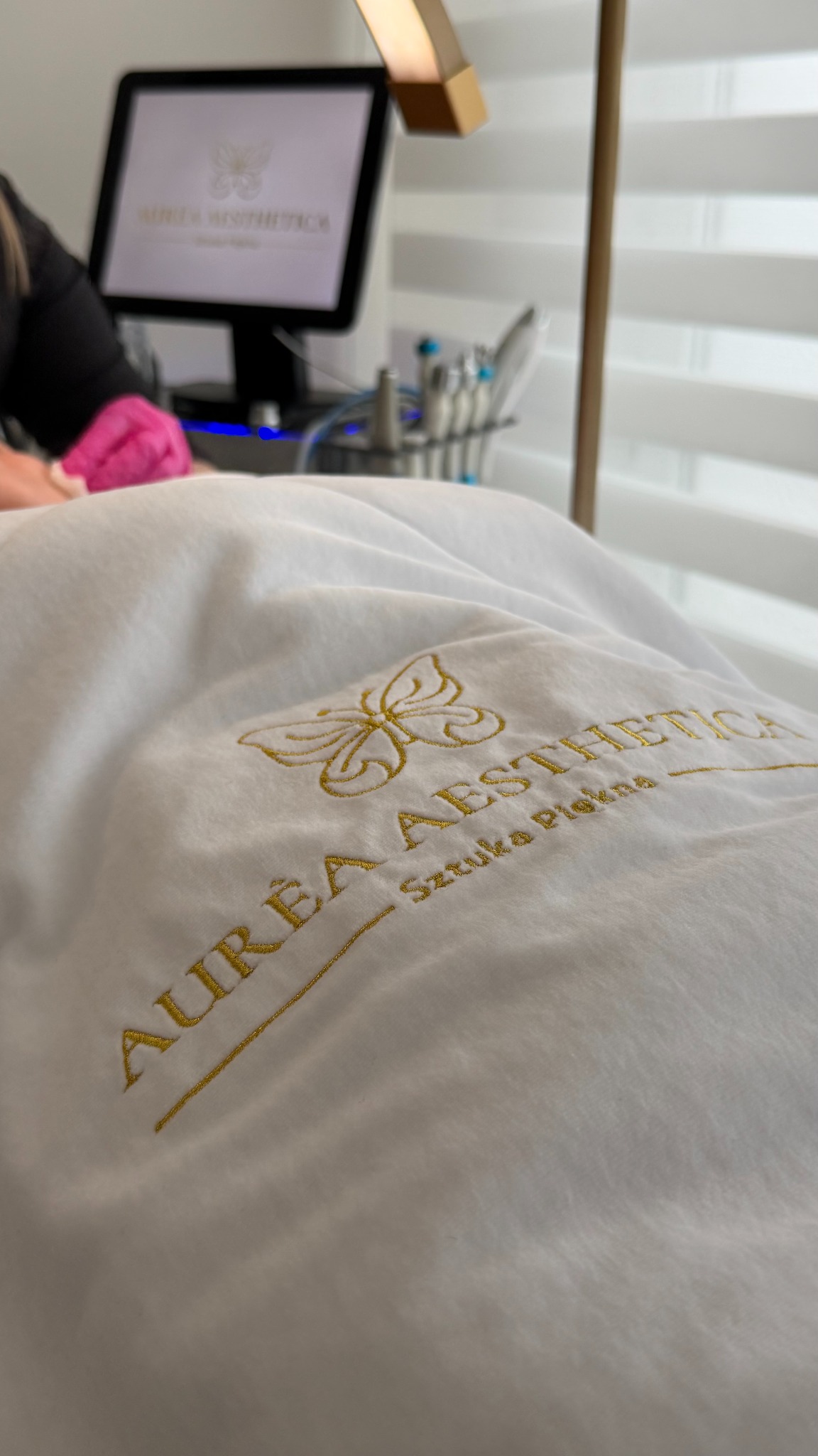 Koc z haftowanym logo salonu kosmetycznego Aurea Aesthetica, w tle monitor z logo i narzędzia kosmetyczne. Ujęcie z perspektywy klienta leżącego na leżance.
